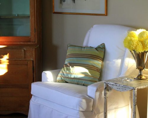 Cozy white armchair with a striped cushion in a sunlit corner.